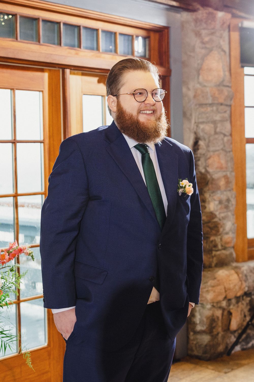 A groom in a navy suit and green tie stands by a window in a rustic stone-walled room.