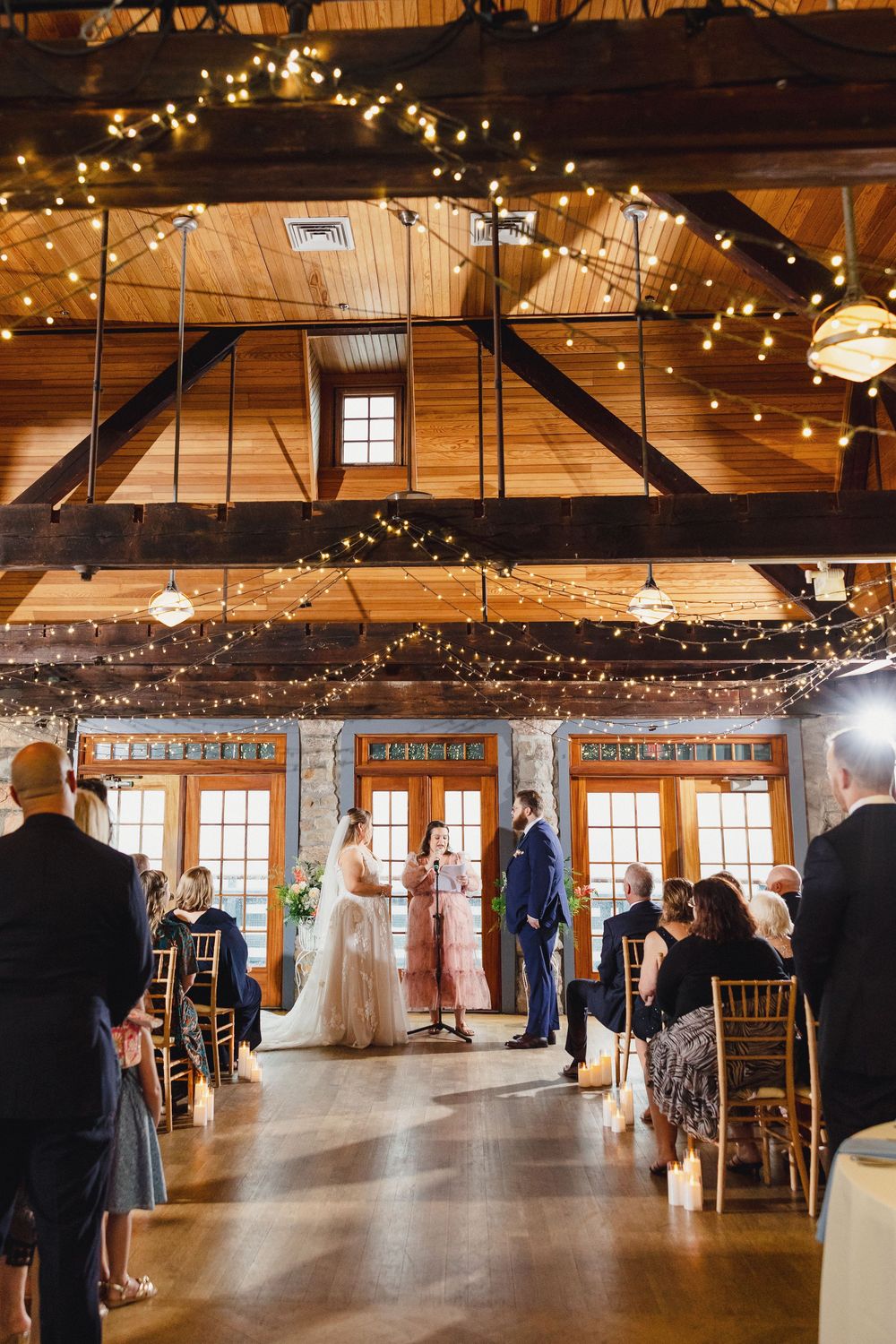Indoor rustic barn wedding ceremony with exposed wooden beams and twinkling string lights illuminating the intimate space.