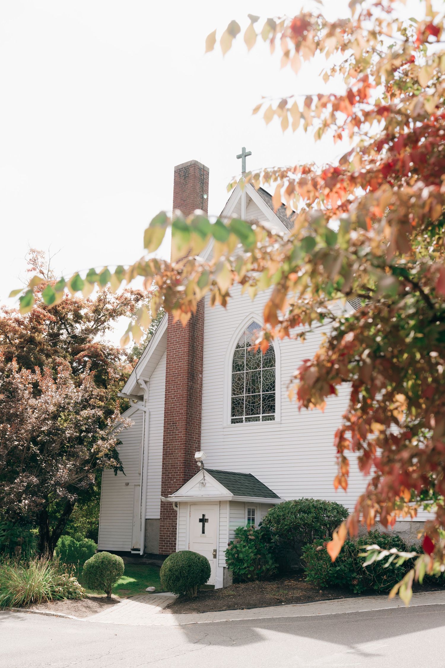 A white church building with brick accents and arched windows surrounded by autumn foliage and landscaping.