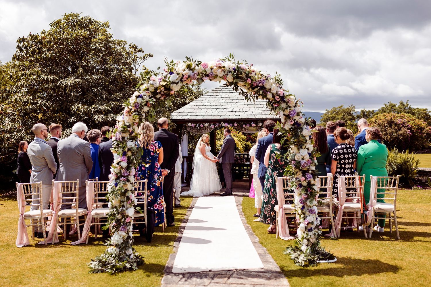 Panoramic view of an outdoor wedding ceremony with floral arch decorations and guests seated on white chairs.