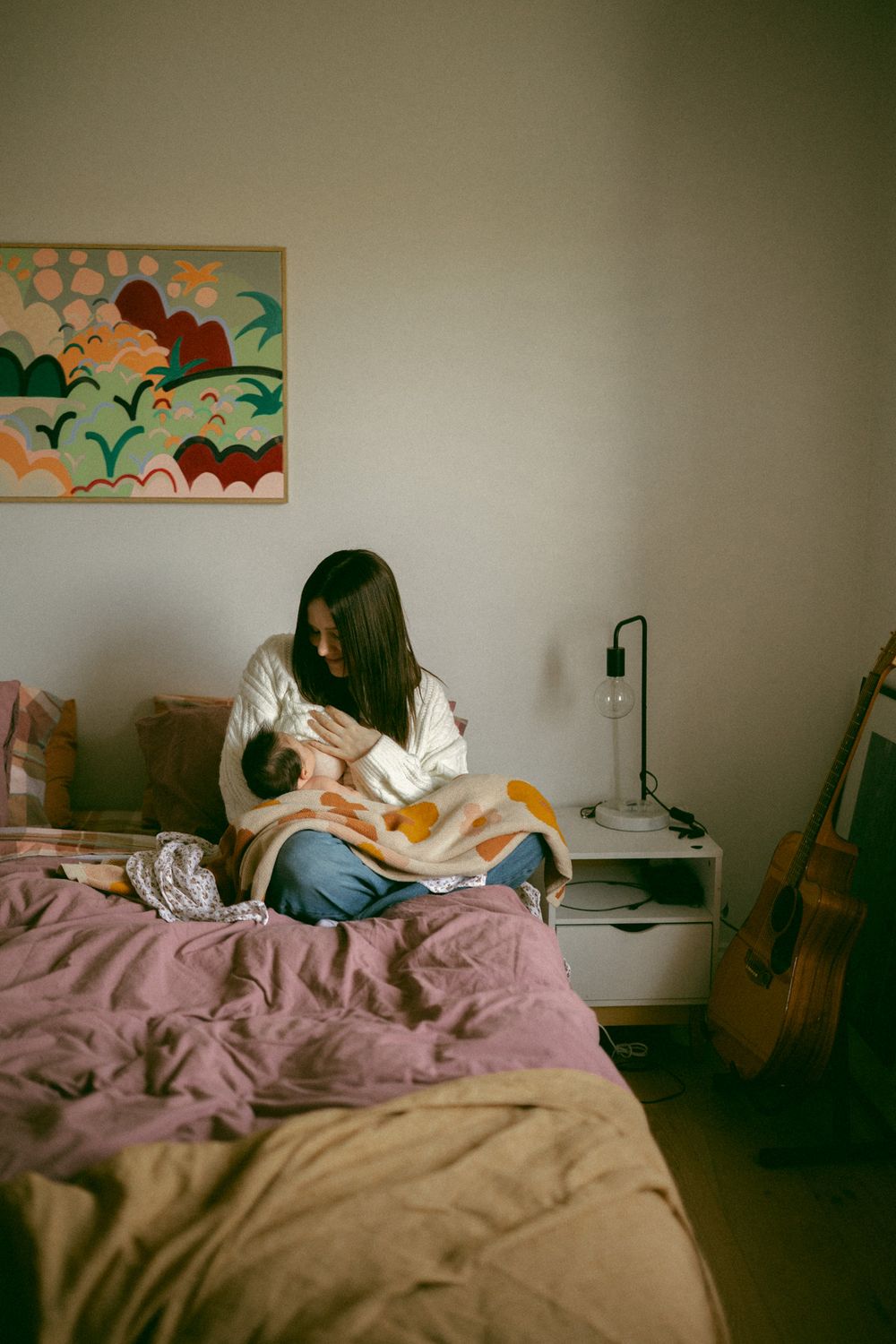 Someone sits cross-legged on an unmade bed with pink sheets, reading in a cozy bedroom with artwork on the wall.