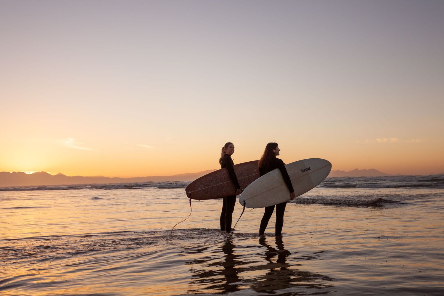 Silhouettes of surfers on a beach at sunset with orange sky and ocean waves.