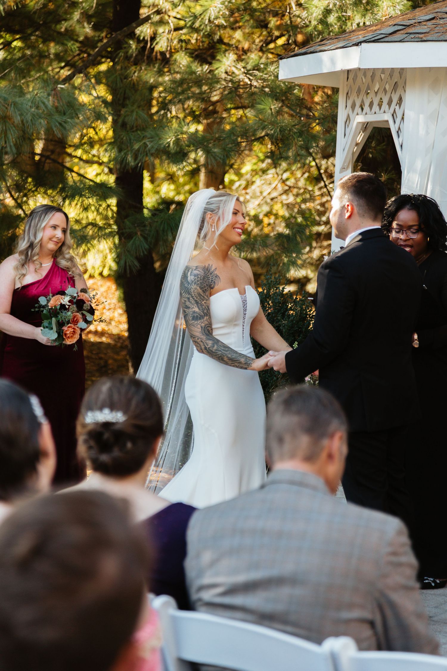 Smiling tattooed bride holding groom's hands during outdoor wedding ceremony near white gazebo.