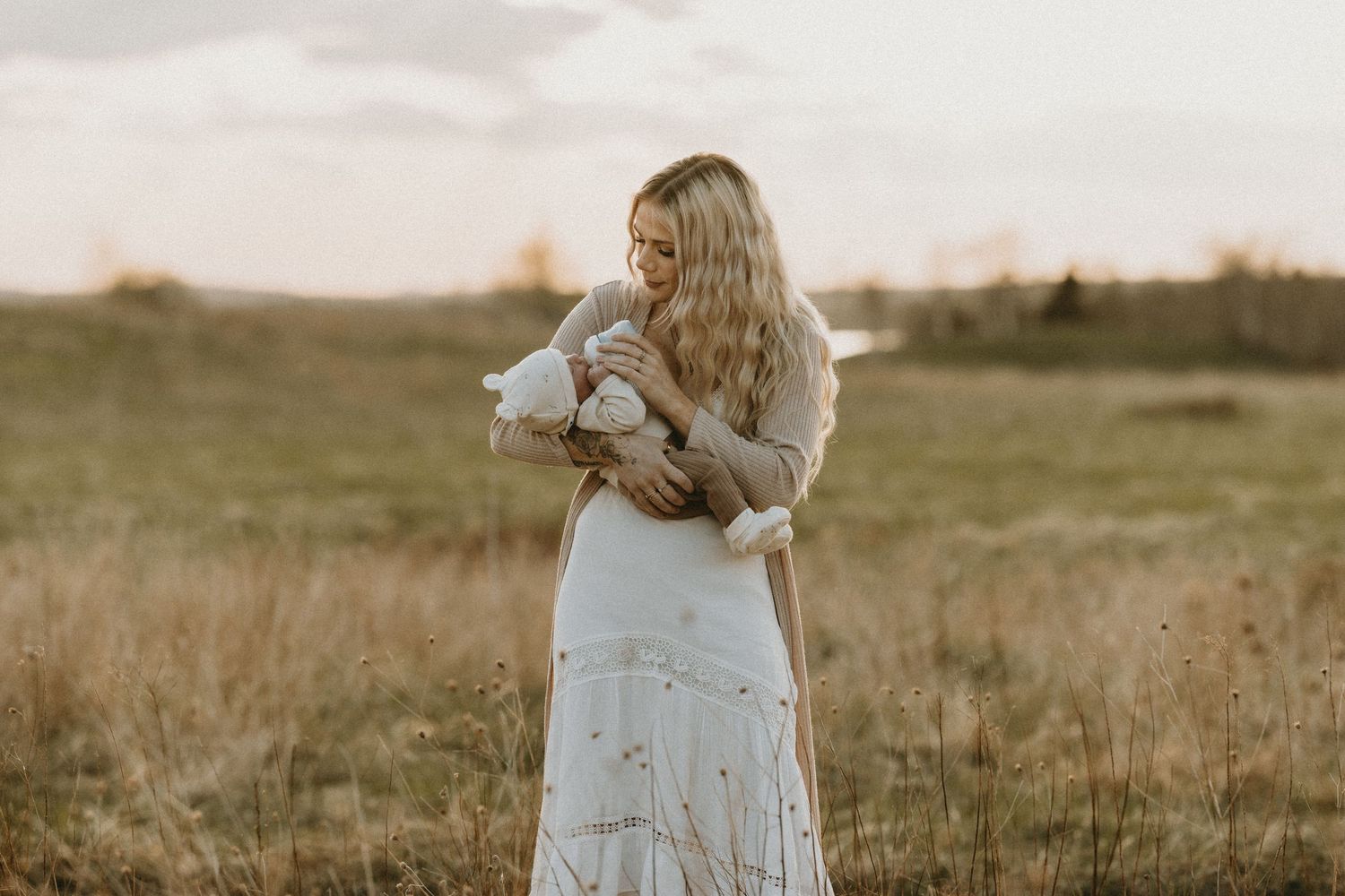 A person in a white dress stands in a golden field during sunset holding flowers.