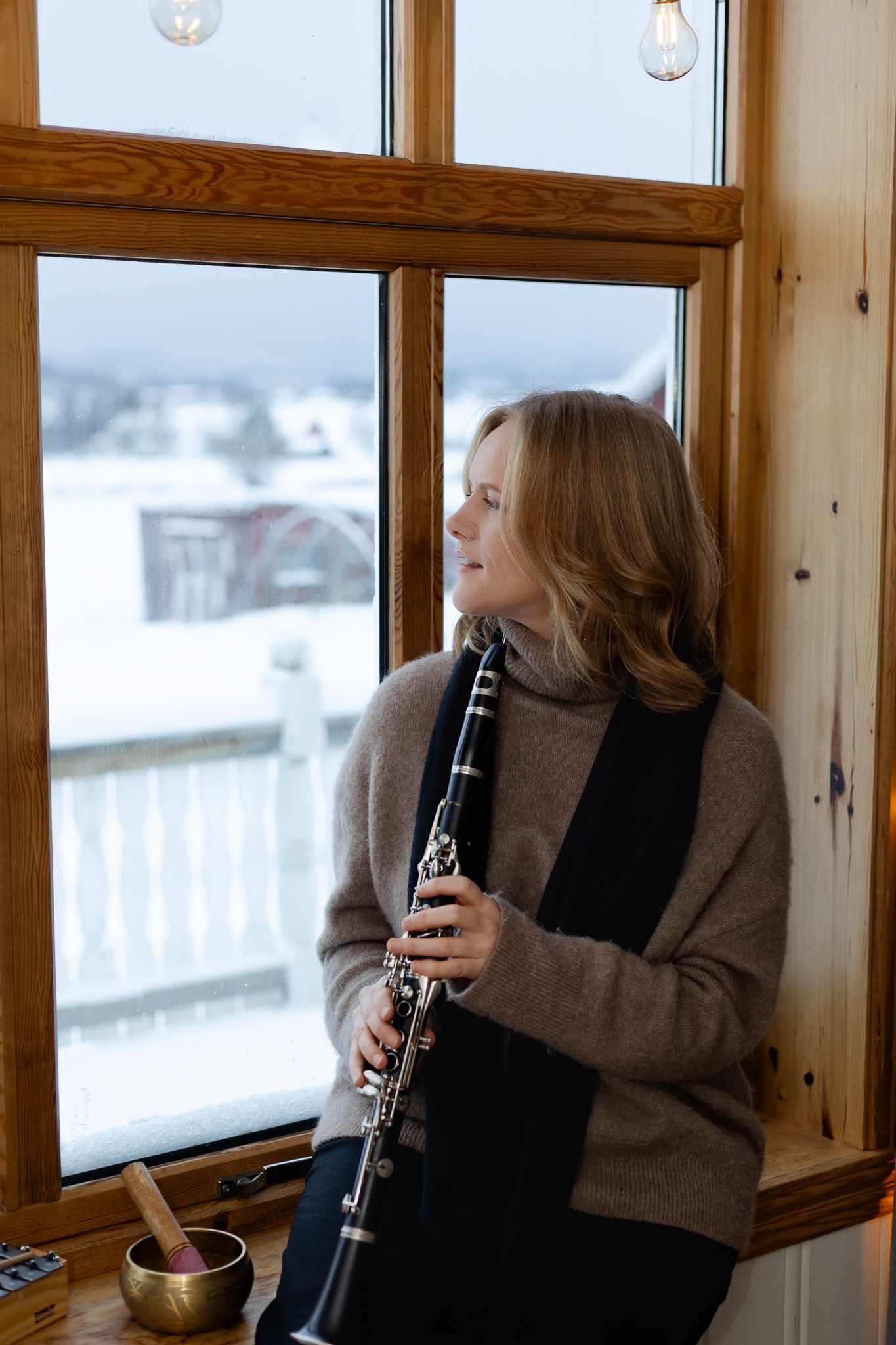 A musician plays clarinet by a row of windows in a cozy wooden room with snowy winter scenery outside.