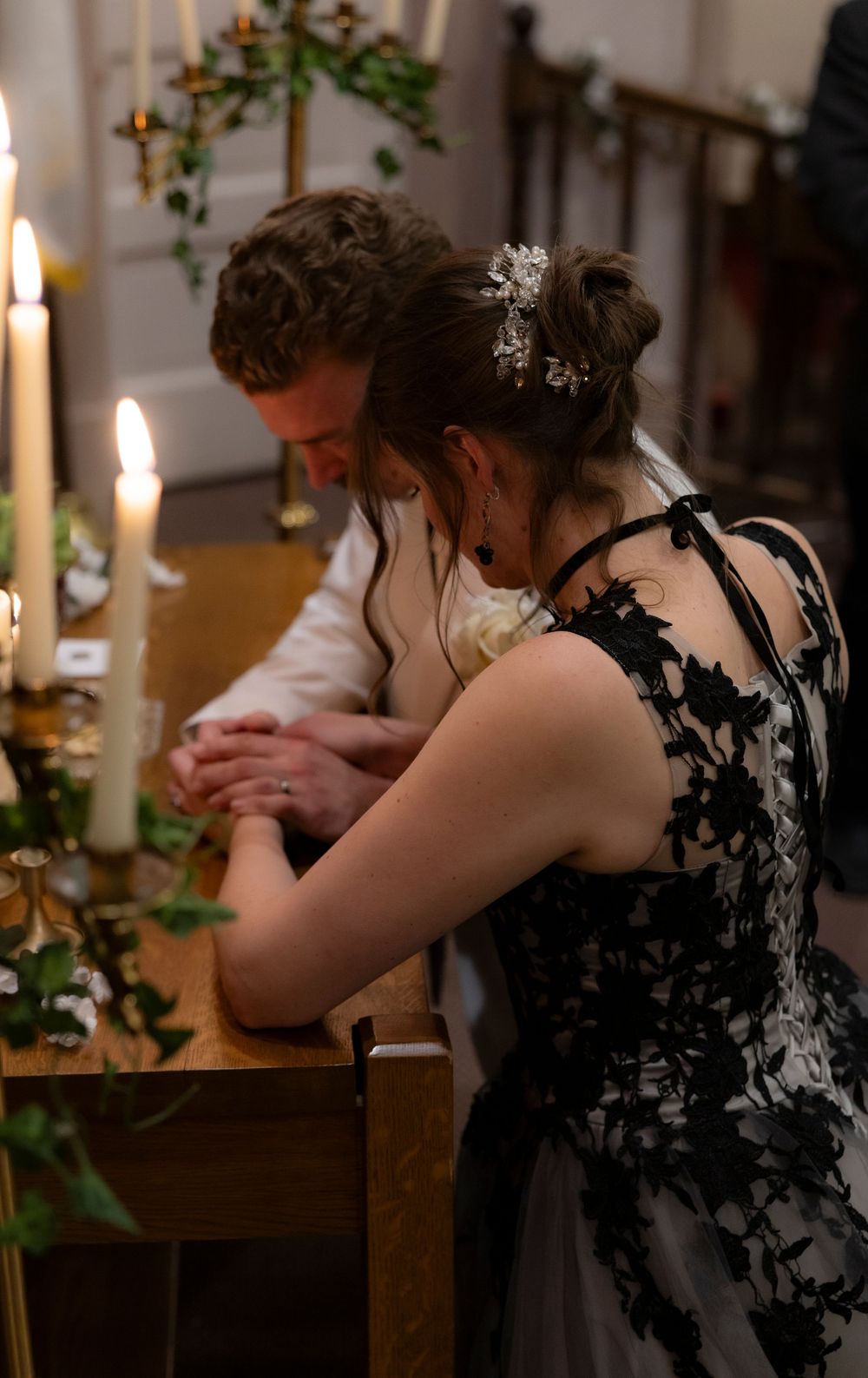 A couple in formal attire sits at a candlelit table with greenery decorations in a romantic setting.