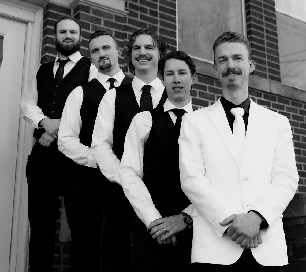 A black and white panoramic photo shows a group of people in formal attire standing together against a brick wall.