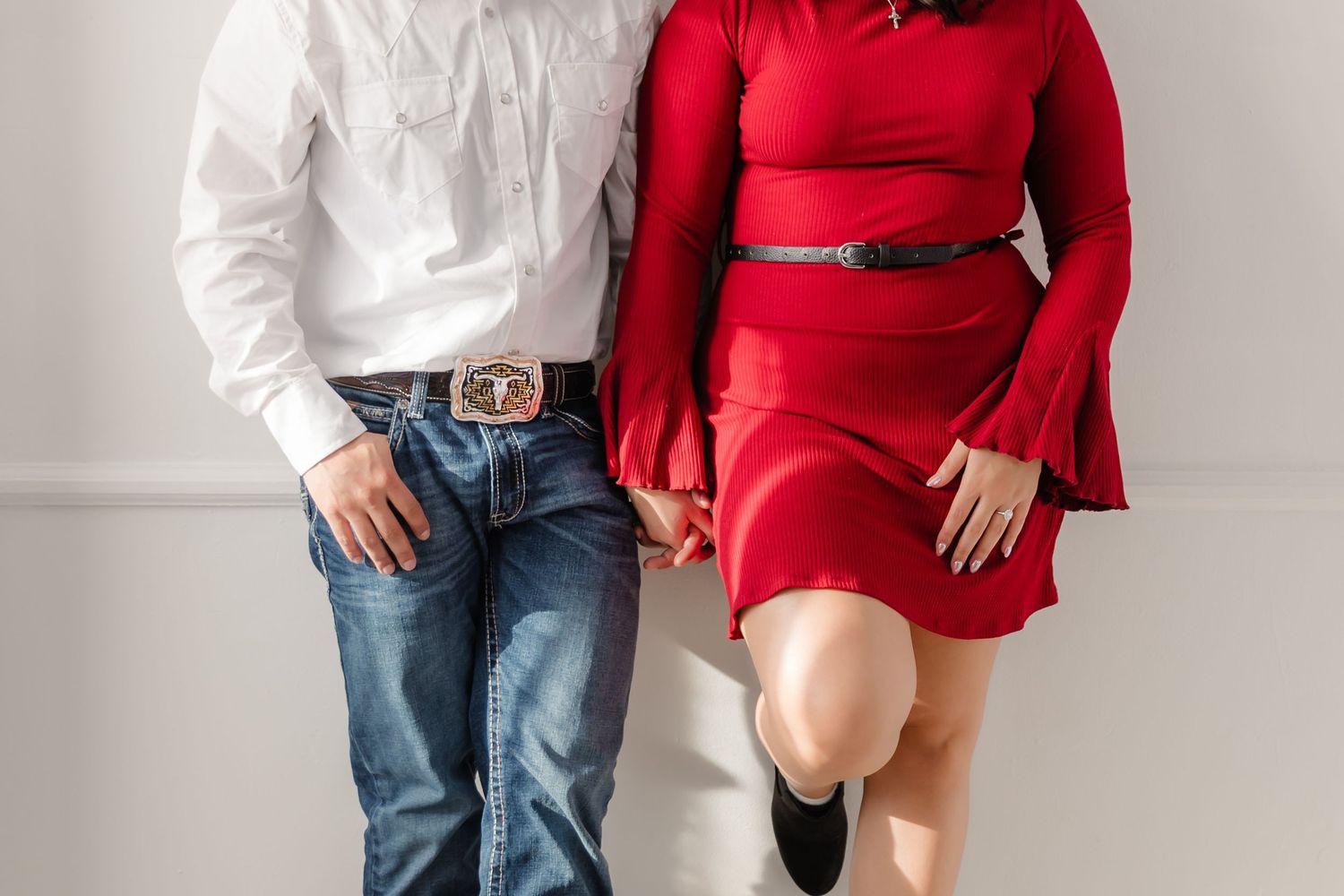 A couple poses against a white wall wearing contrasting outfits of denim with white shirt and a red two-piece dress.