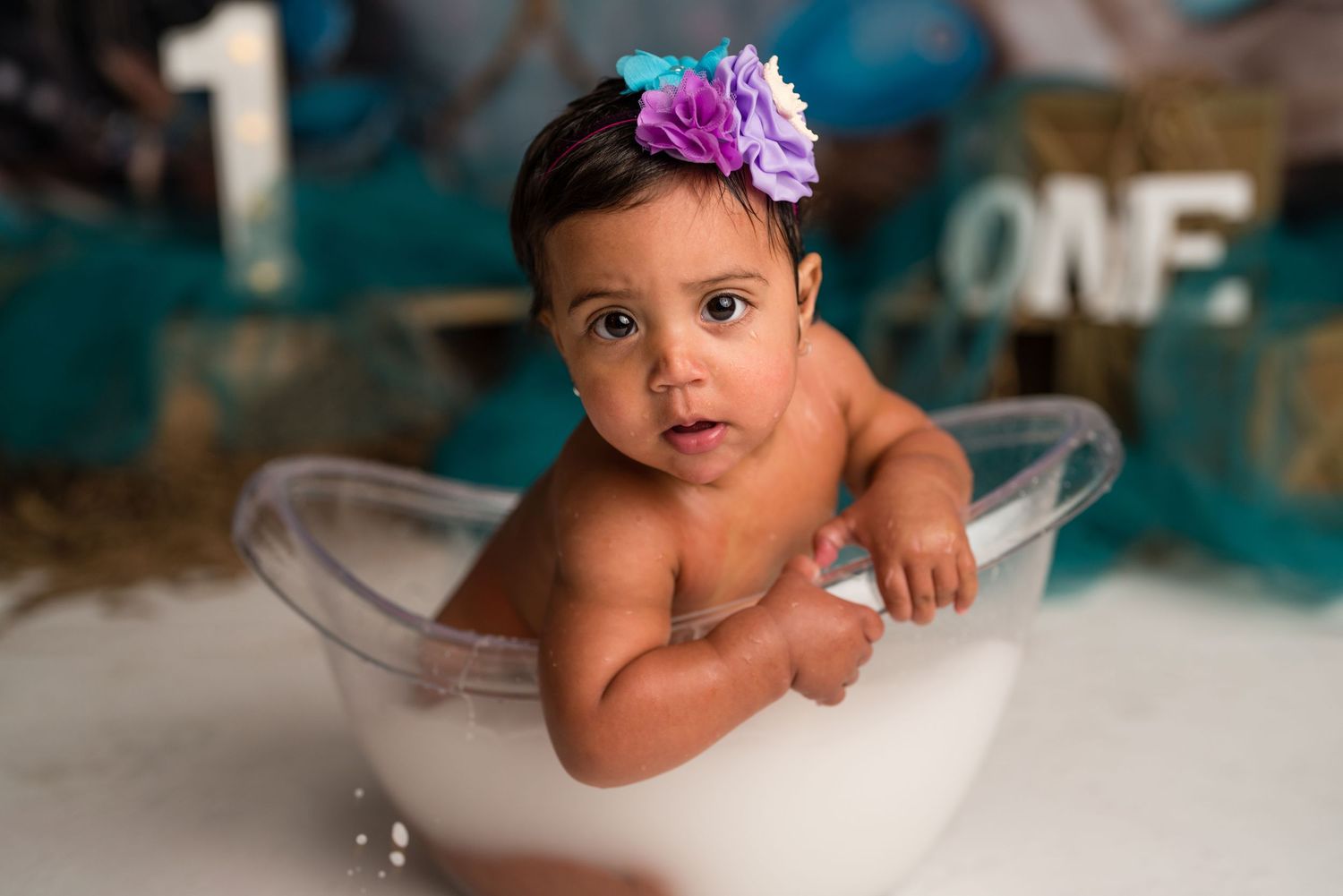 A baby in a white dress sits in a bathtub during a professional photo session with turquoise backdrop.