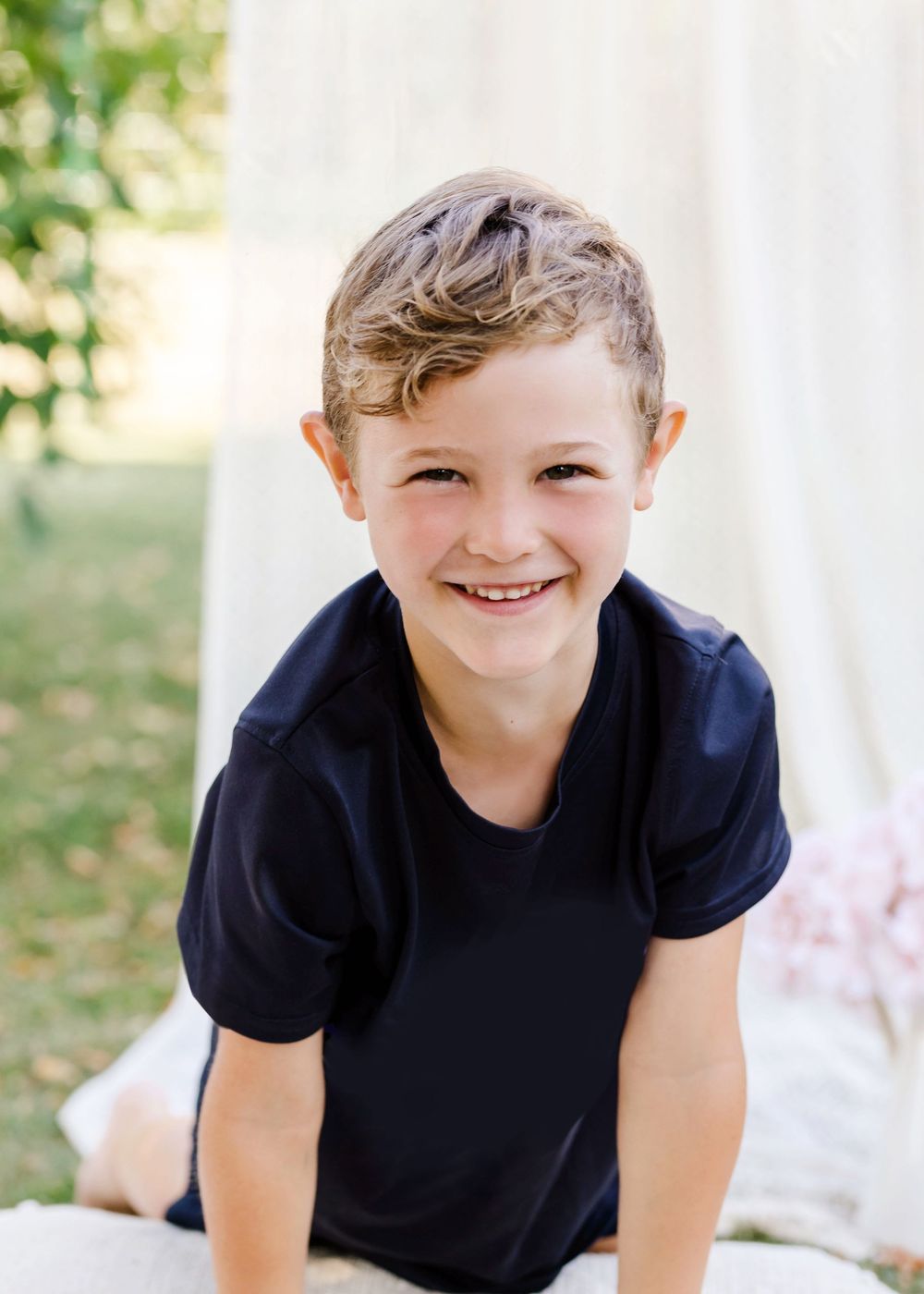 Child wearing navy blue t-shirt smiles brightly while sitting outdoors against white background in natural lighting.