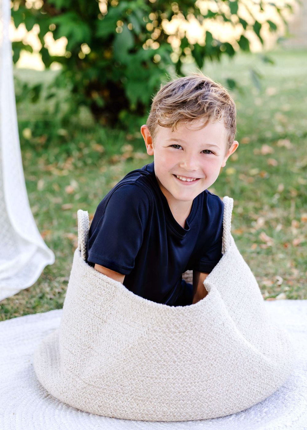 Child in navy shirt playfully peeks through curved white cutout while sitting on grass outdoors.