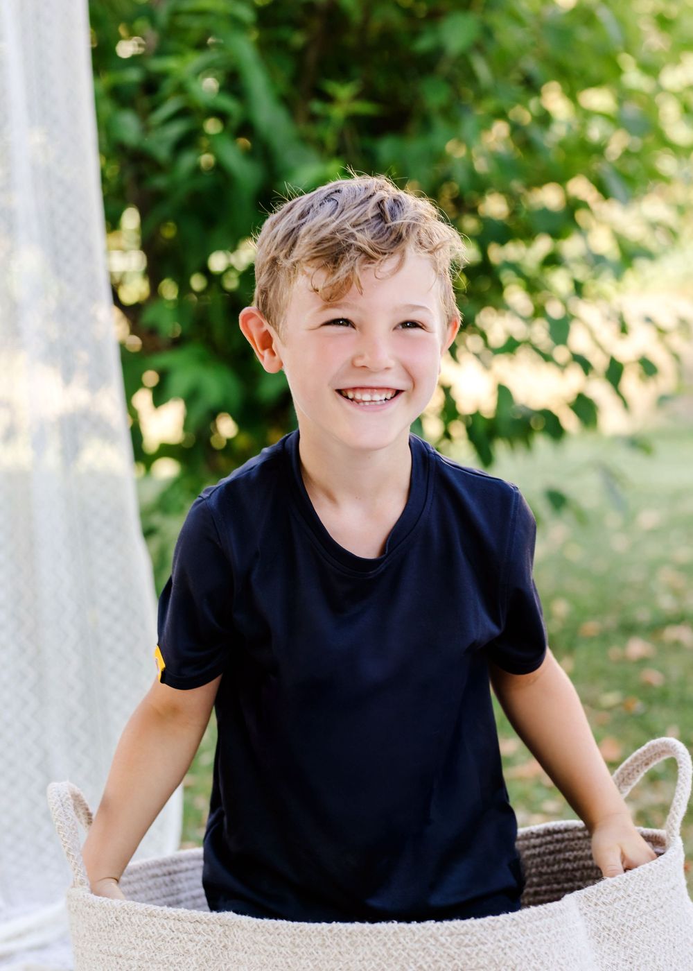 Child relaxing on white blanket outdoors with green foliage in background.