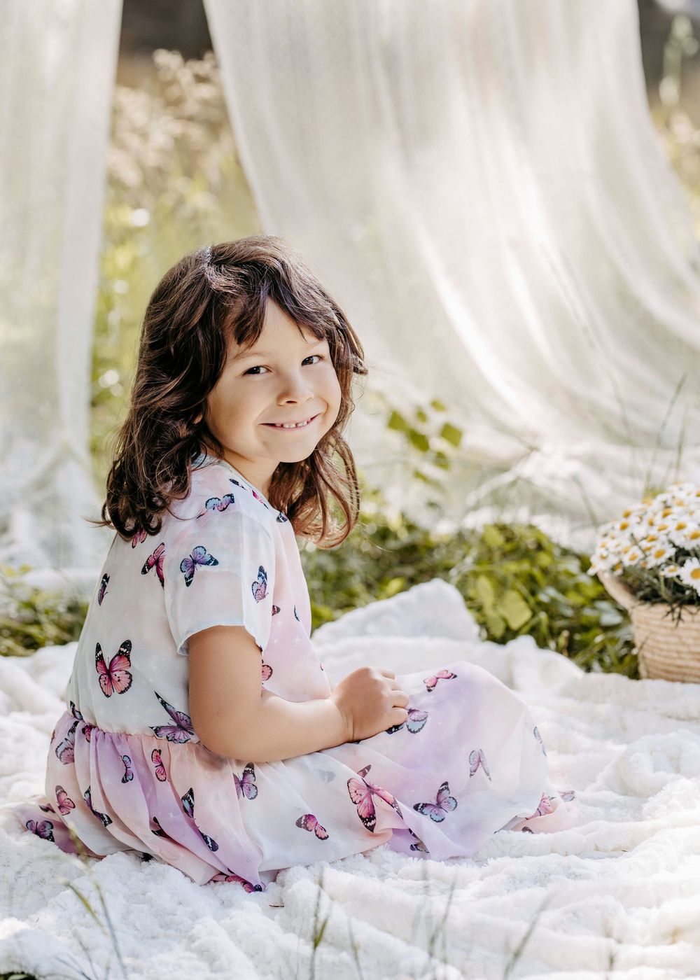 A child in a butterfly print dress lies on white fabric outdoors surrounded by delicate wildflowers and sunlight.