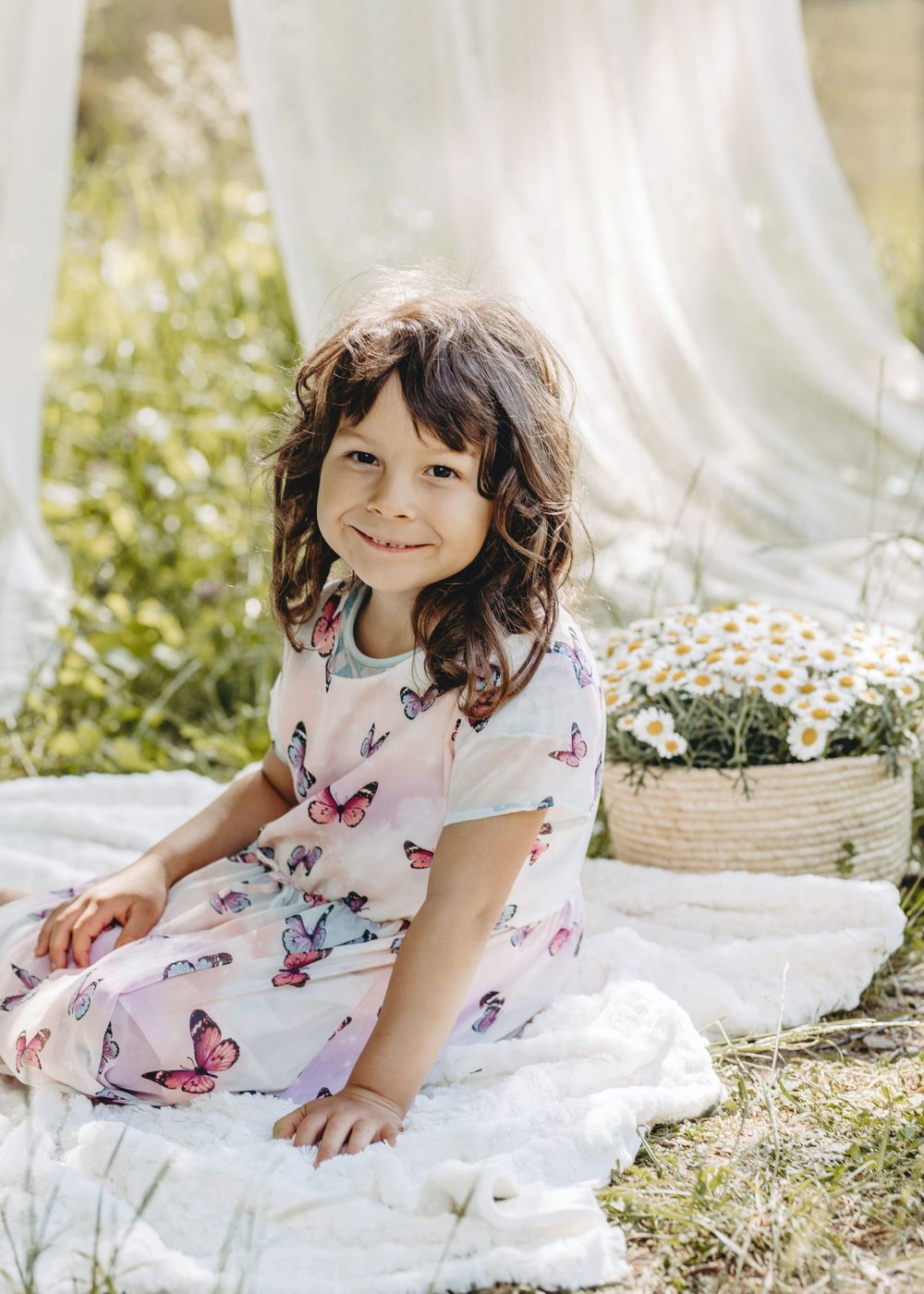 A child in a white butterfly dress poses on a blanket outdoors during a sunny photoshoot.