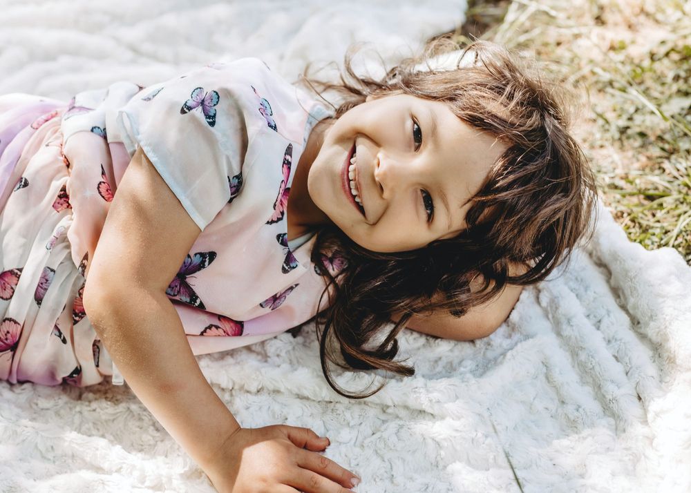 A child lying on a white blanket outdoors wearing a butterfly print dress in a dreamy garden setting.