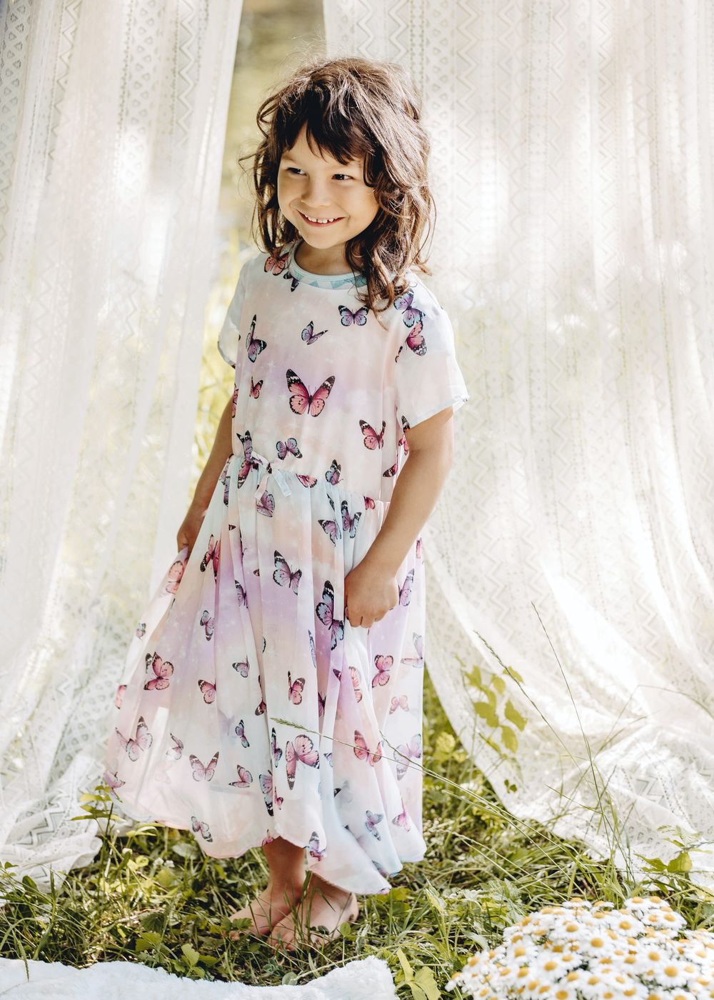 A child in a butterfly-patterned dress plays near sheer white curtains in a whimsical outdoor setting.