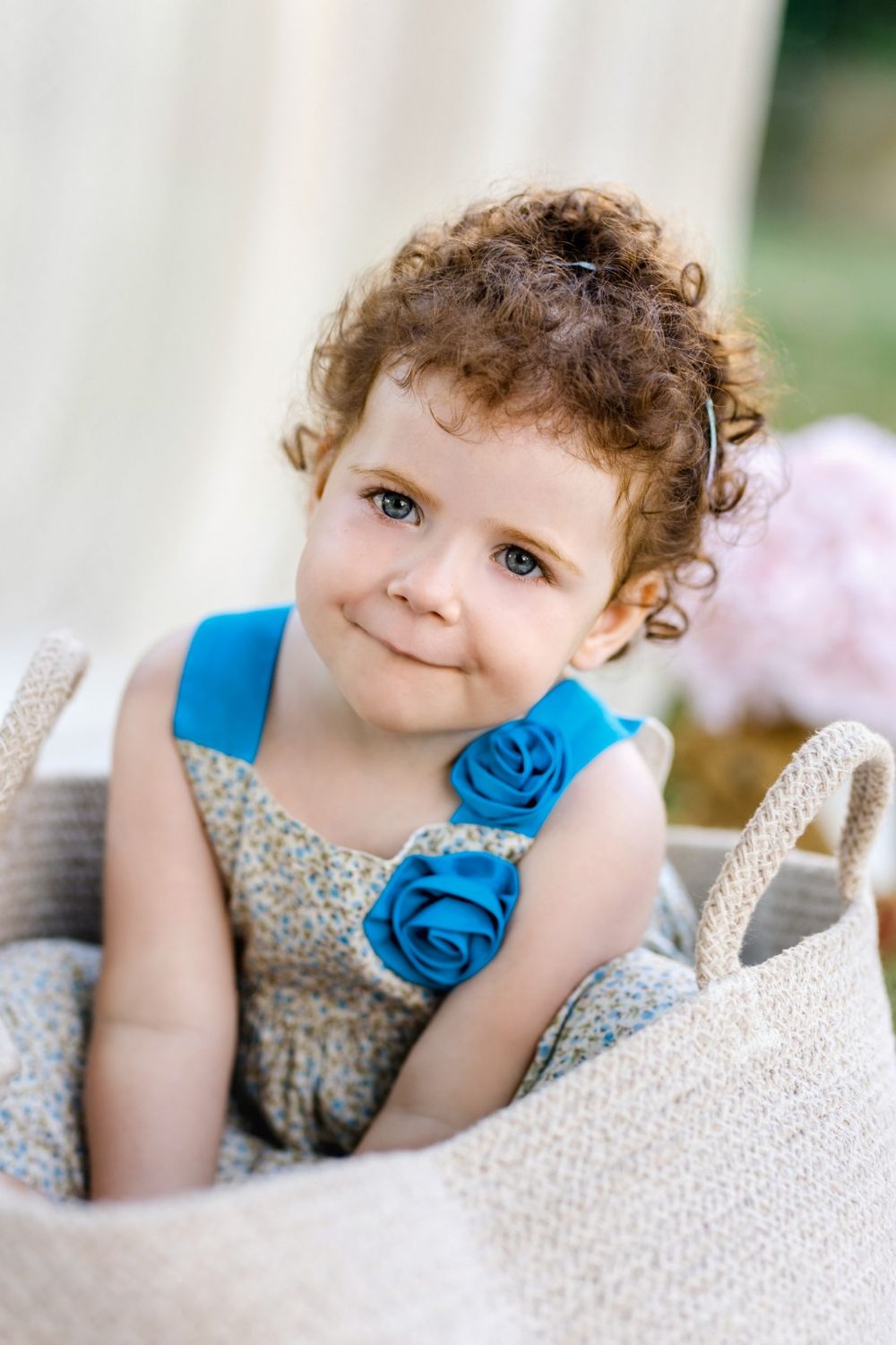 A child with curly hair wearing a floral dress and blue flower accessory rests on white textured fabric.