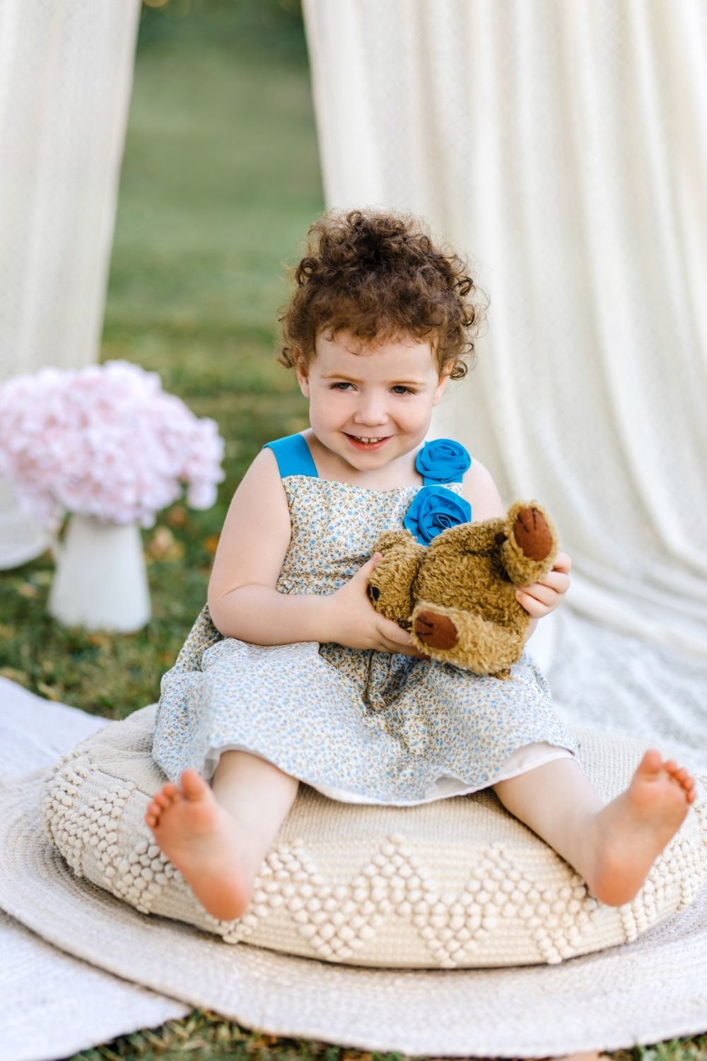 A child in a polka dot dress sits on a white cushion holding a teddy bear during an outdoor photo session.