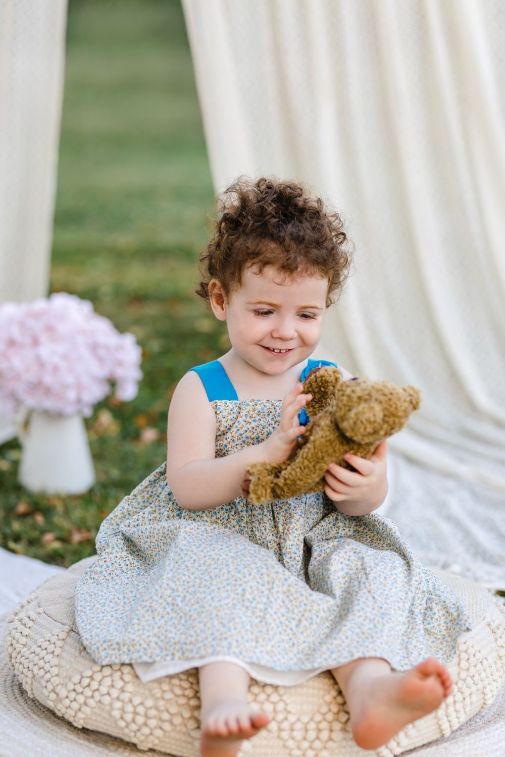 A young child in a floral dress sits on a white blanket outdoors playing with a teddy bear.