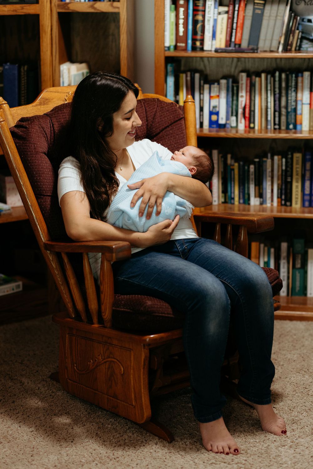 Peaceful moment in home library with mother and baby resting in wooden rocking chair. IN-home newborn photos.