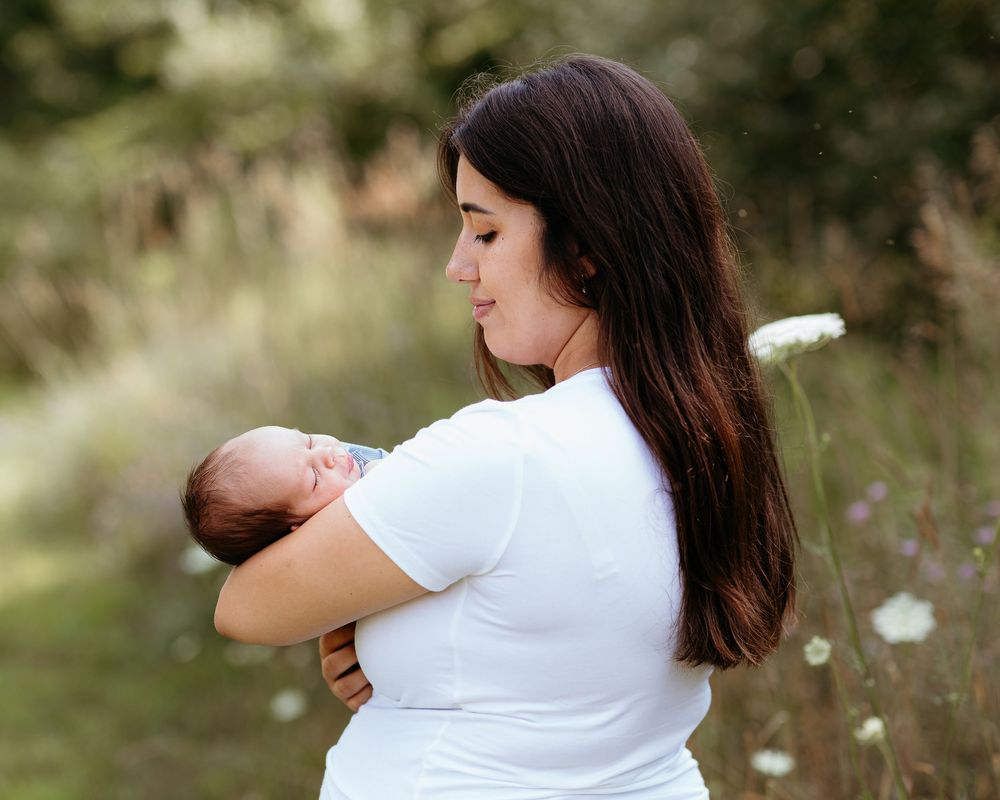 Mother in white shirt holding baby outdoors in natural summer meadow setting.