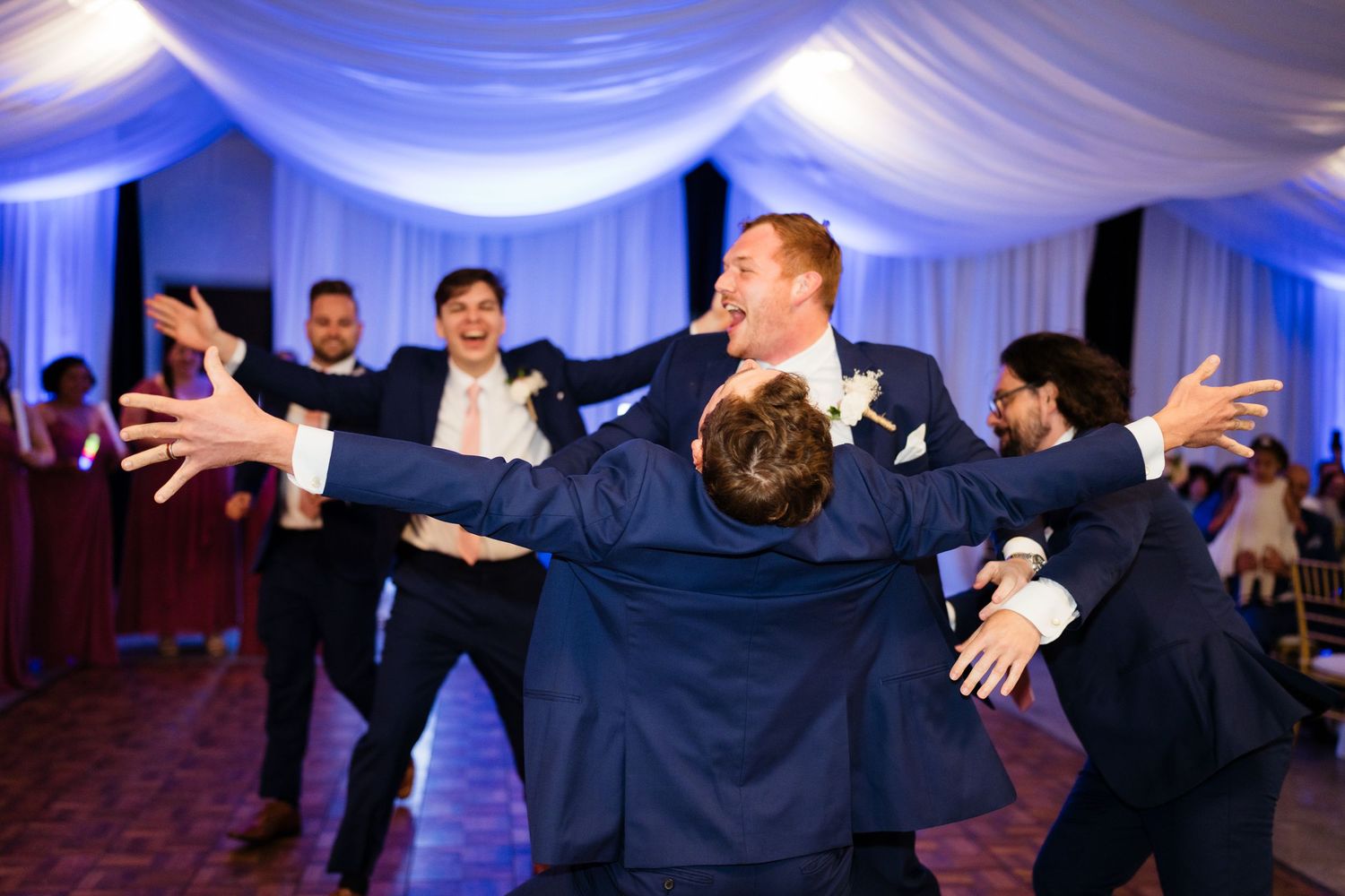 Wedding guests dance with arms raised in celebration under elegant blue-lit draped fabric ceiling at reception venue.