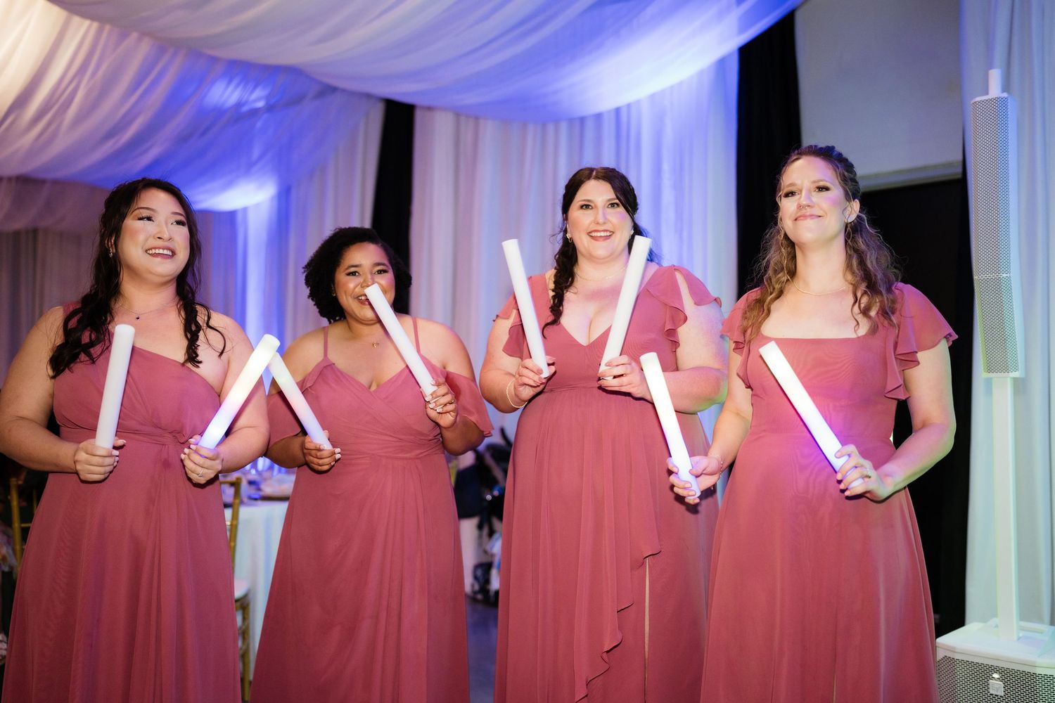 Bridesmaids in matching burgundy dresses pose together at a formal wedding celebration with white draped backdrop.