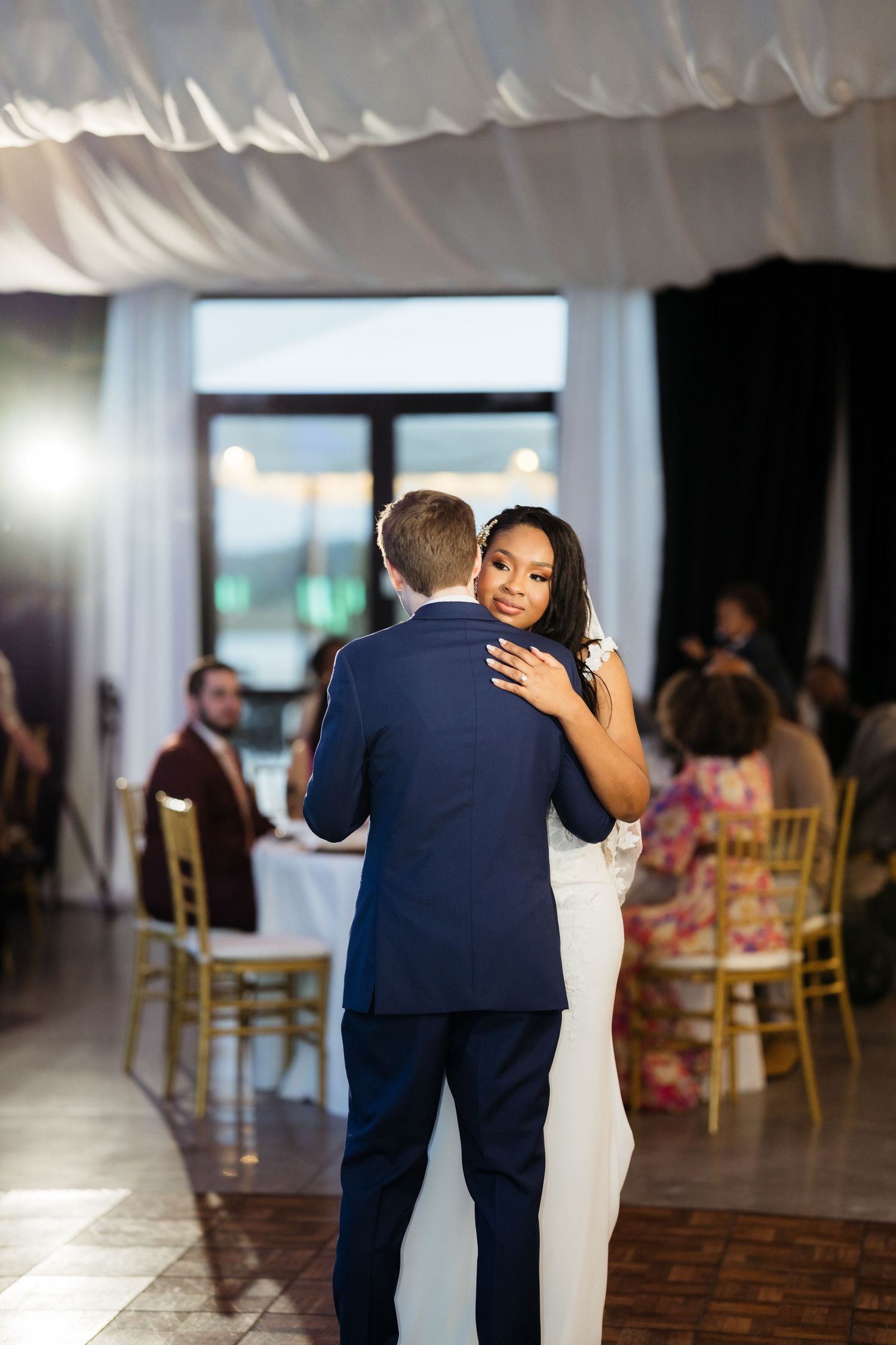 A couple shares an intimate dance moment at a wedding reception with guests seated at tables in the background.