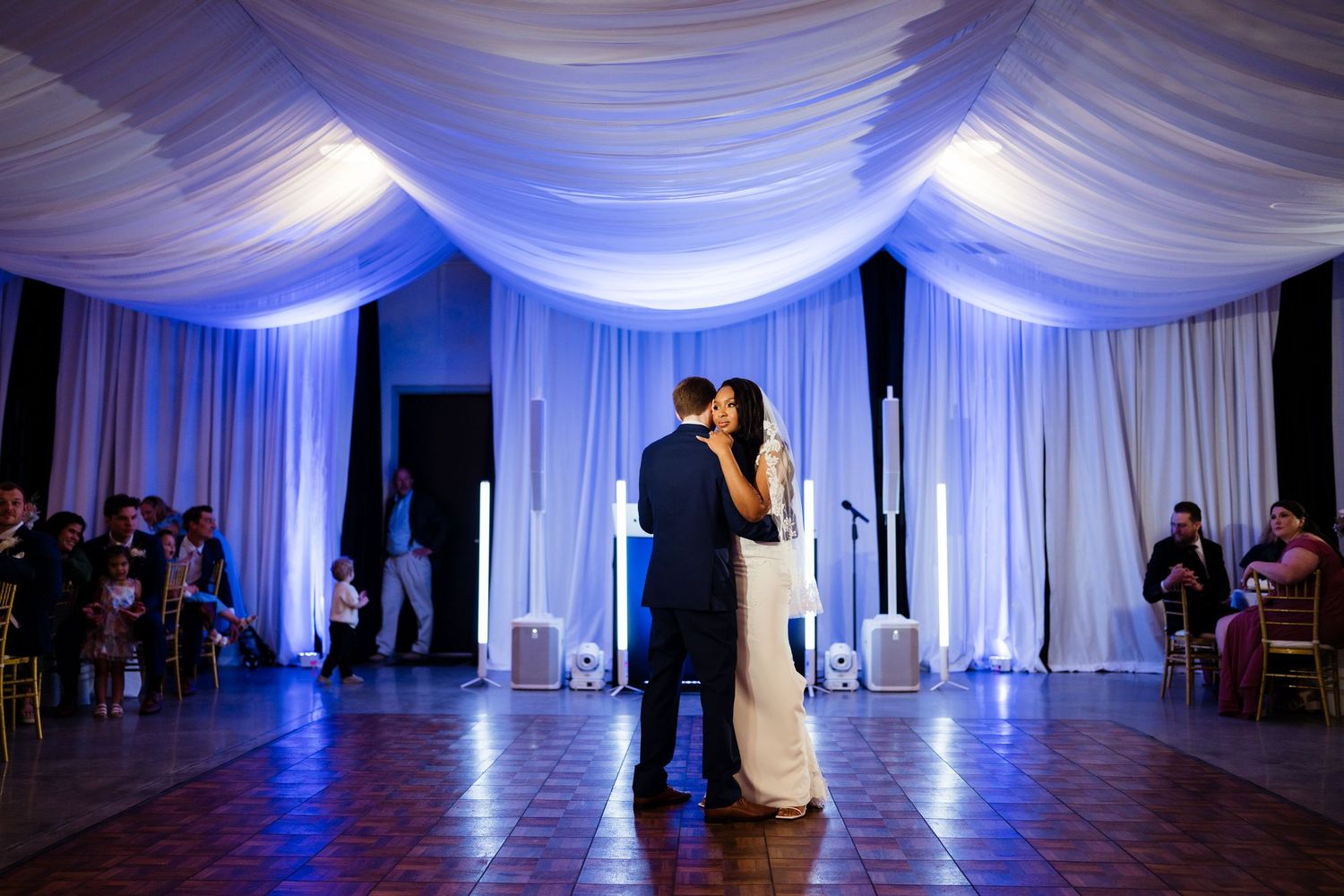 Wedding reception venue with elegant white fabric draping and dramatic blue uplighting creating a romantic atmosphere.