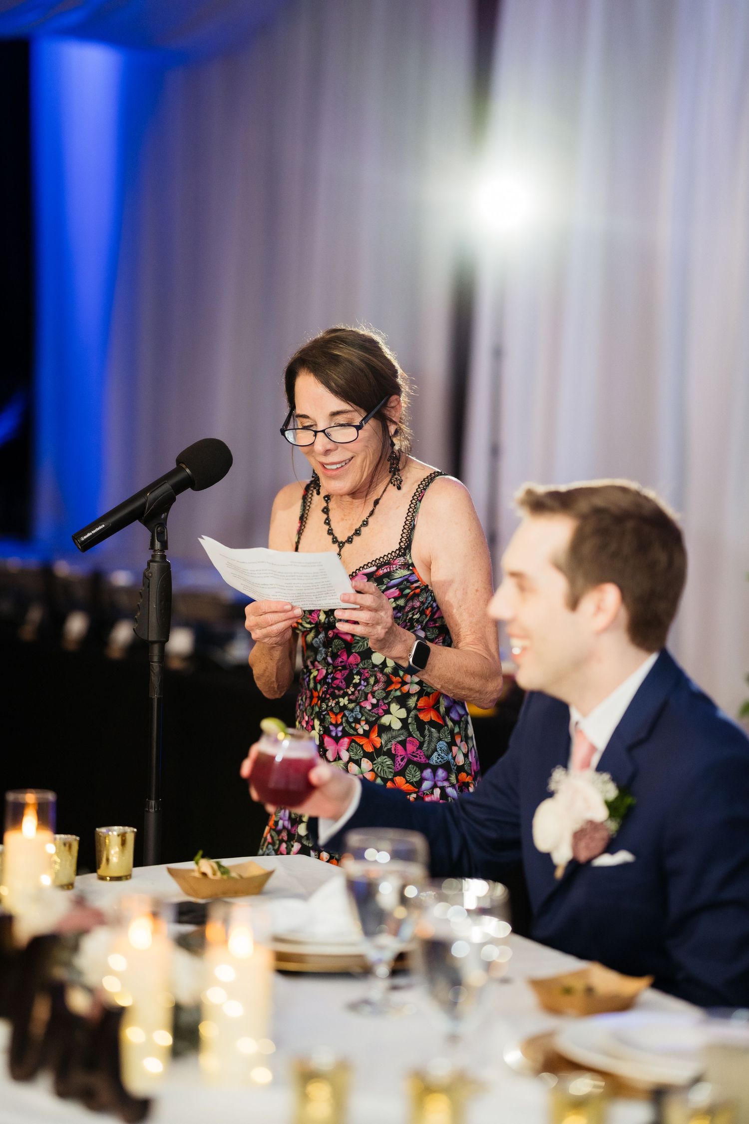 A speaker at a formal dinner event reads from papers at a podium with blue lighting in the background.