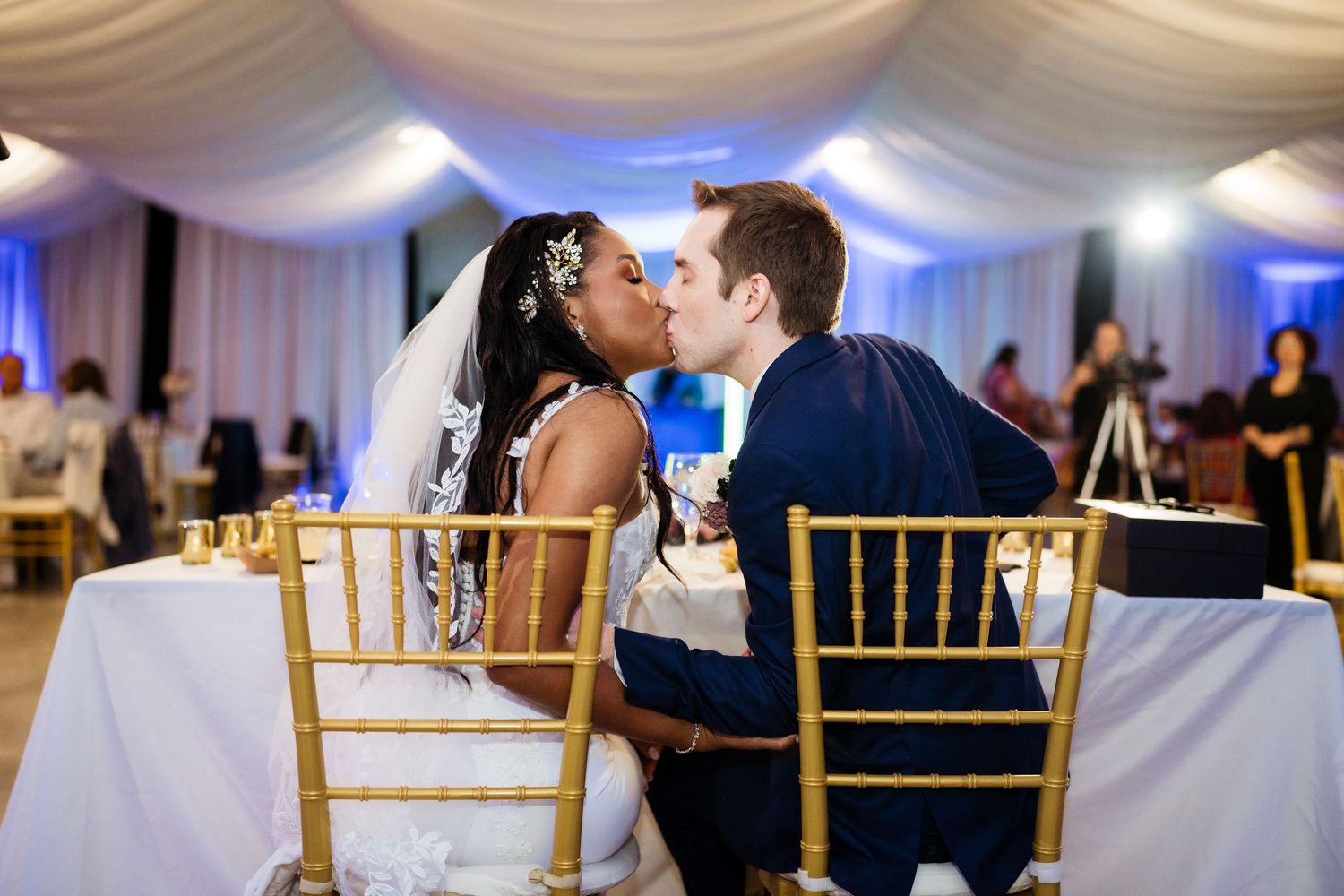 A couple shares a romantic kiss at their wedding reception while seated at gold chiavari chairs.