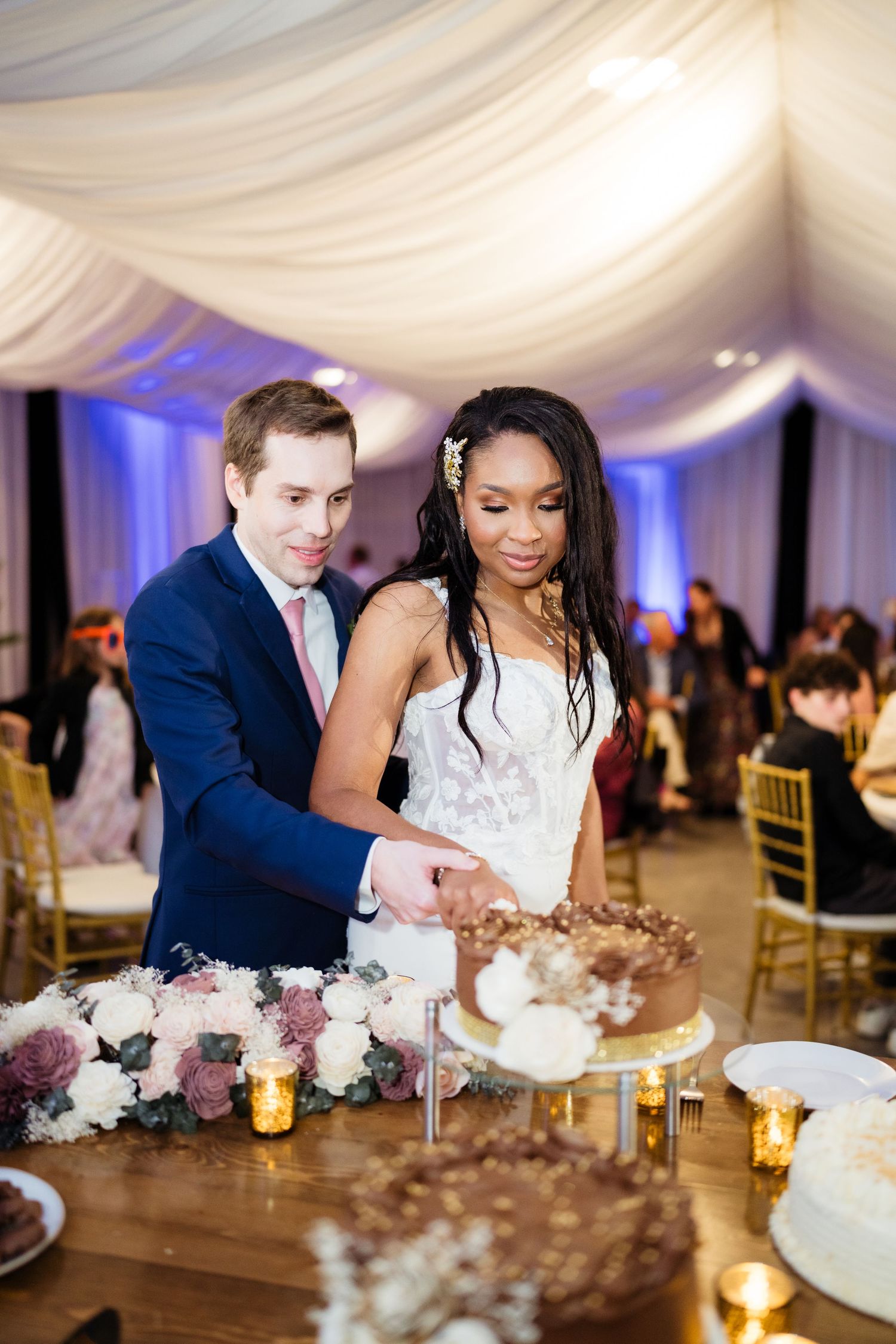 Couple cutting their elegant white wedding cake at a reception decorated with candlelight and floral centerpieces.
