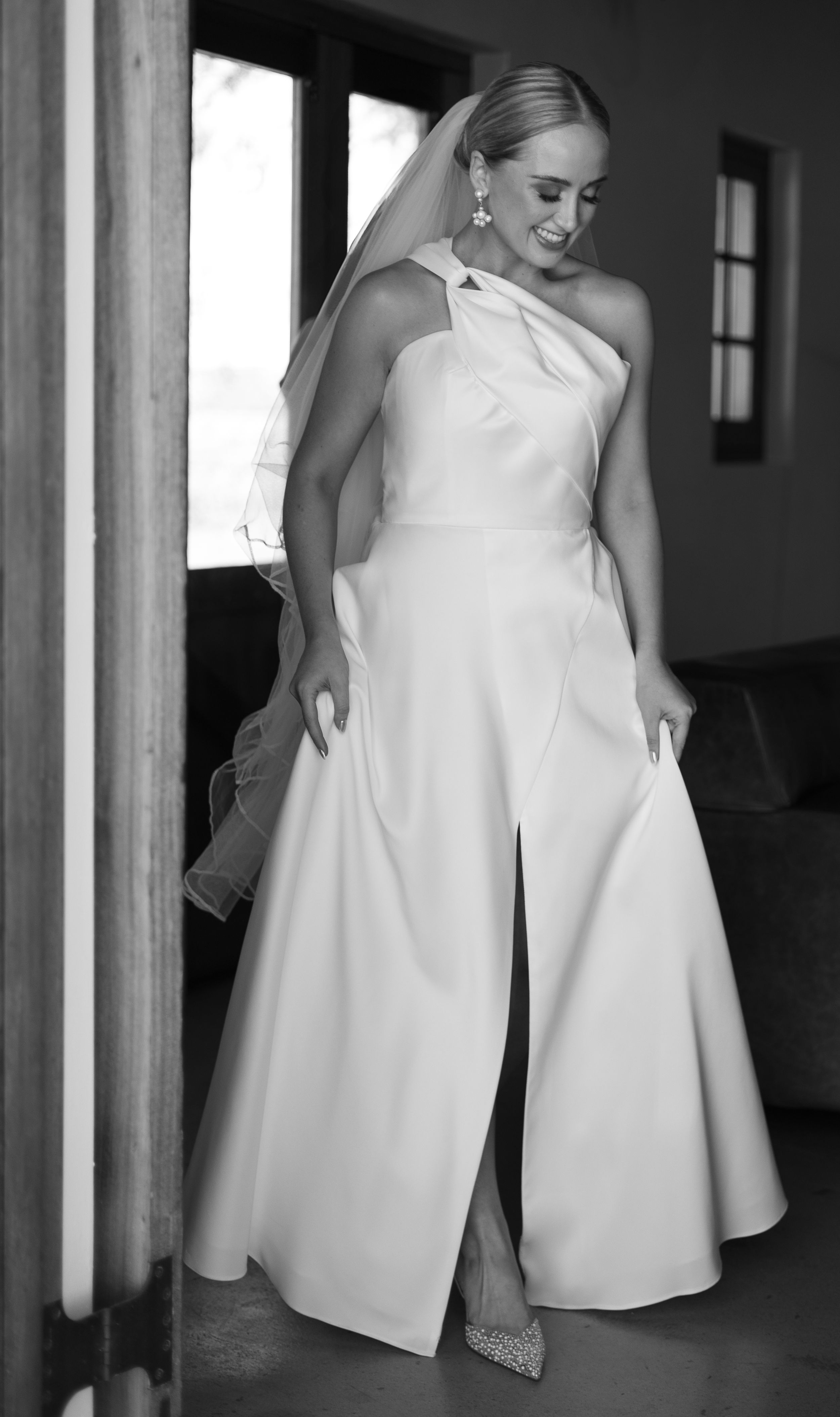 Elegant bride in minimalist one-shoulder white wedding jumpsuit with veil in black and white photo.