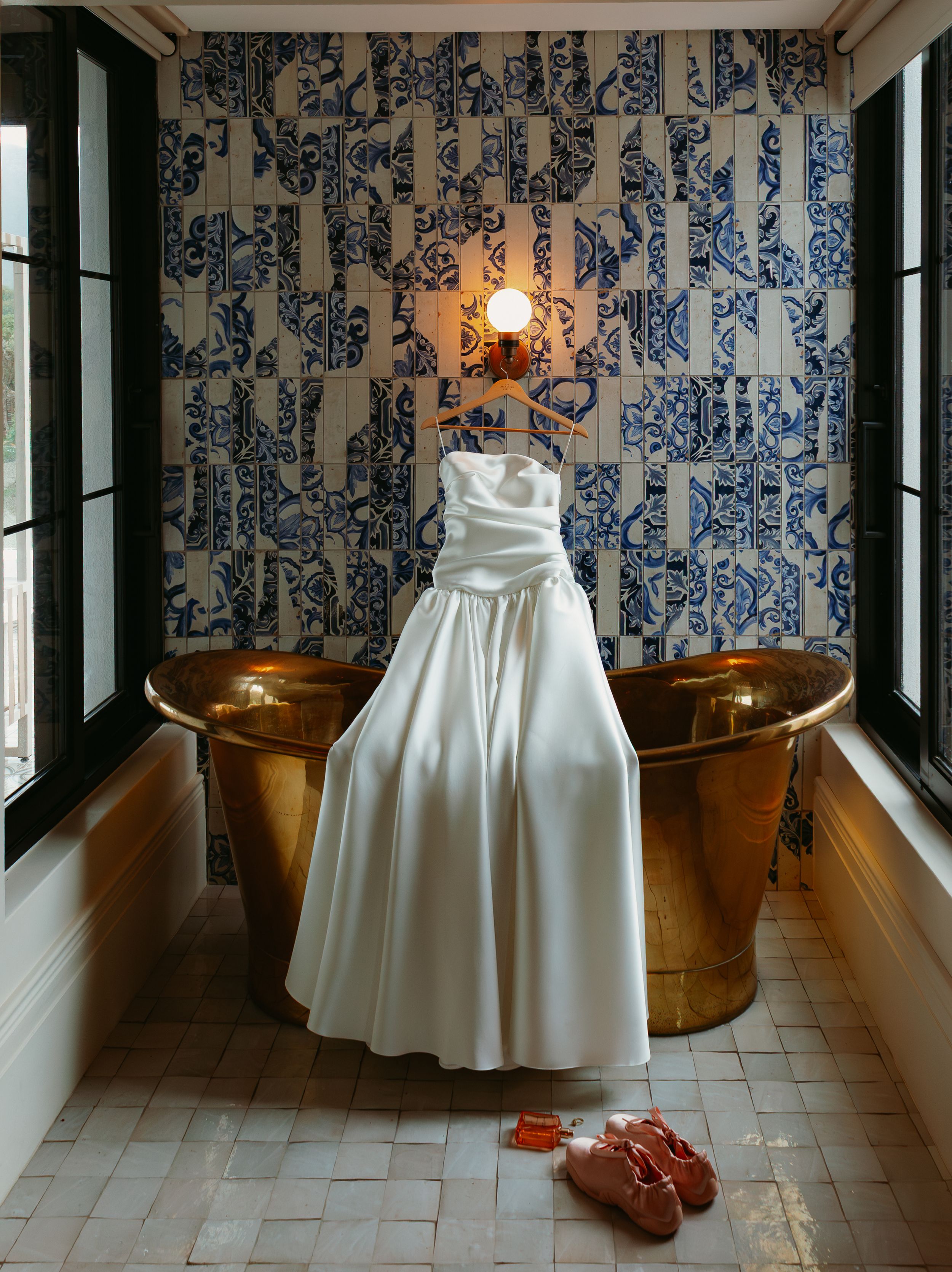 White strapless satin wedding gown hanging above a copper bathtub with blue patterned tile wall.