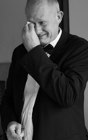 Emotional man in black tuxedo wiping tears at wedding ceremony in black and white photo.