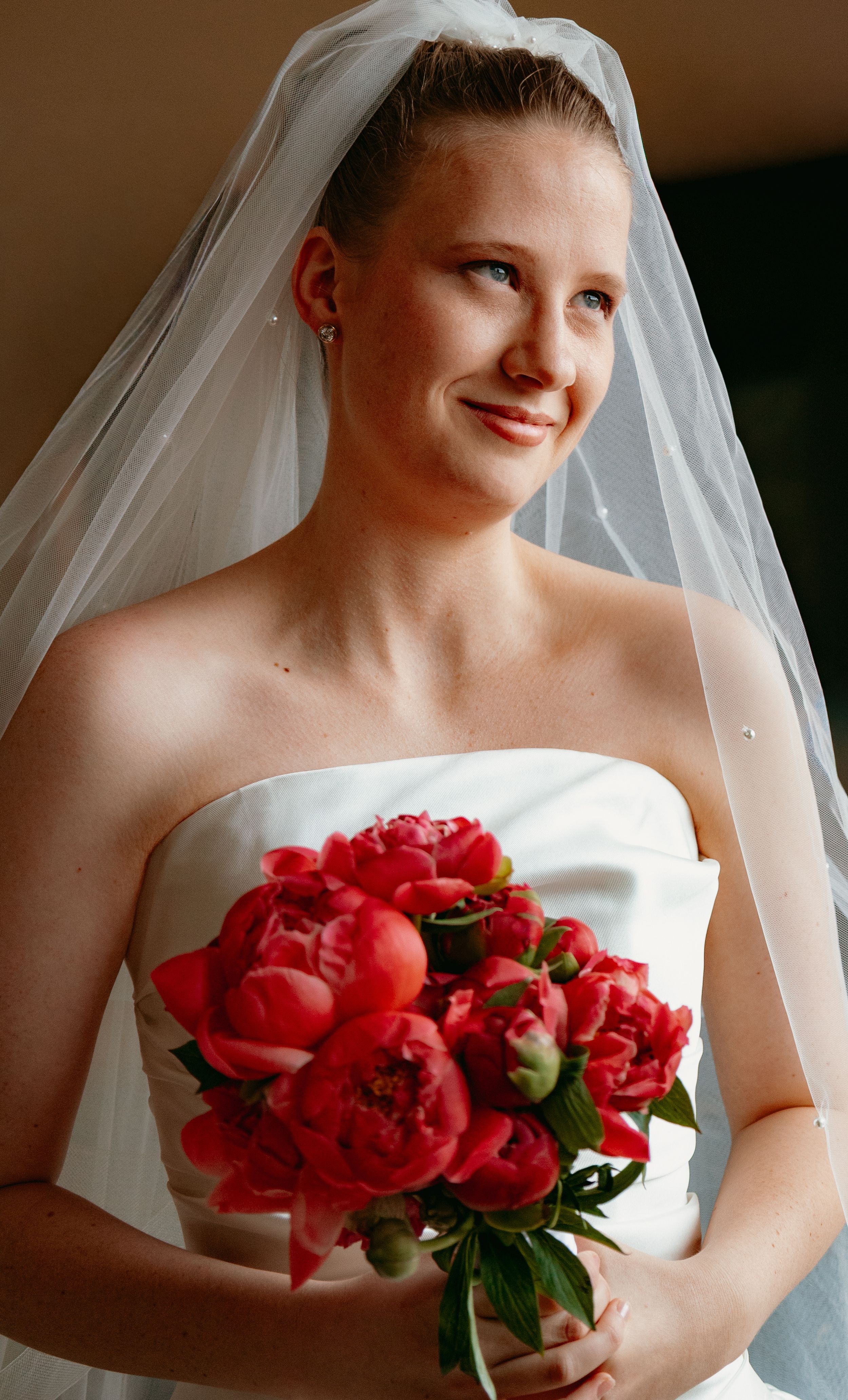 Bride in strapless white gown holding vibrant red peony bouquet wearing veil smiling softly.
