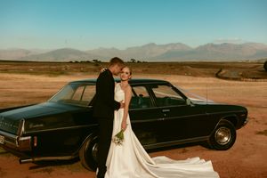 Bride and groom posing beside vintage black car with mountain landscape backdrop at golden hour.