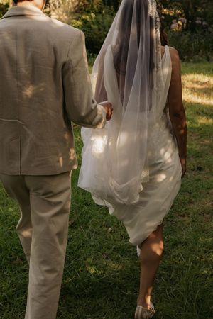 Bride and groom walking hand in hand through green grass, bride wearing short white dress and flowing veil in golden light.