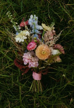 Colorful wildflower wedding bouquet with dahlias, delphiniums, and mixed blooms wrapped in pink ribbon on green grass.