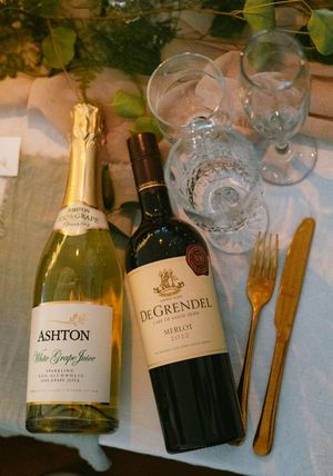 Ashton white grape juice and De Grendel Merlot wine bottles on wedding reception table with gold cutlery and glasses.