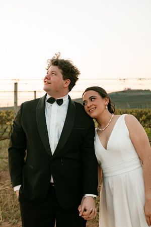 Happy bride and groom holding hands in vineyard at sunset, groom in black tuxedo with bow tie.
