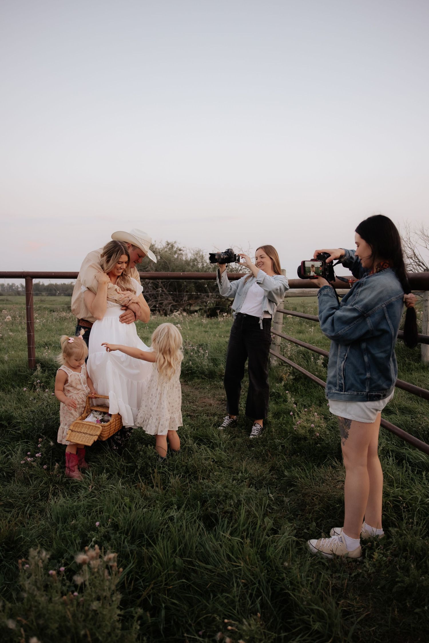 Photographers capture a family photo session in a rustic outdoor setting during golden hour at a countryside fence.
