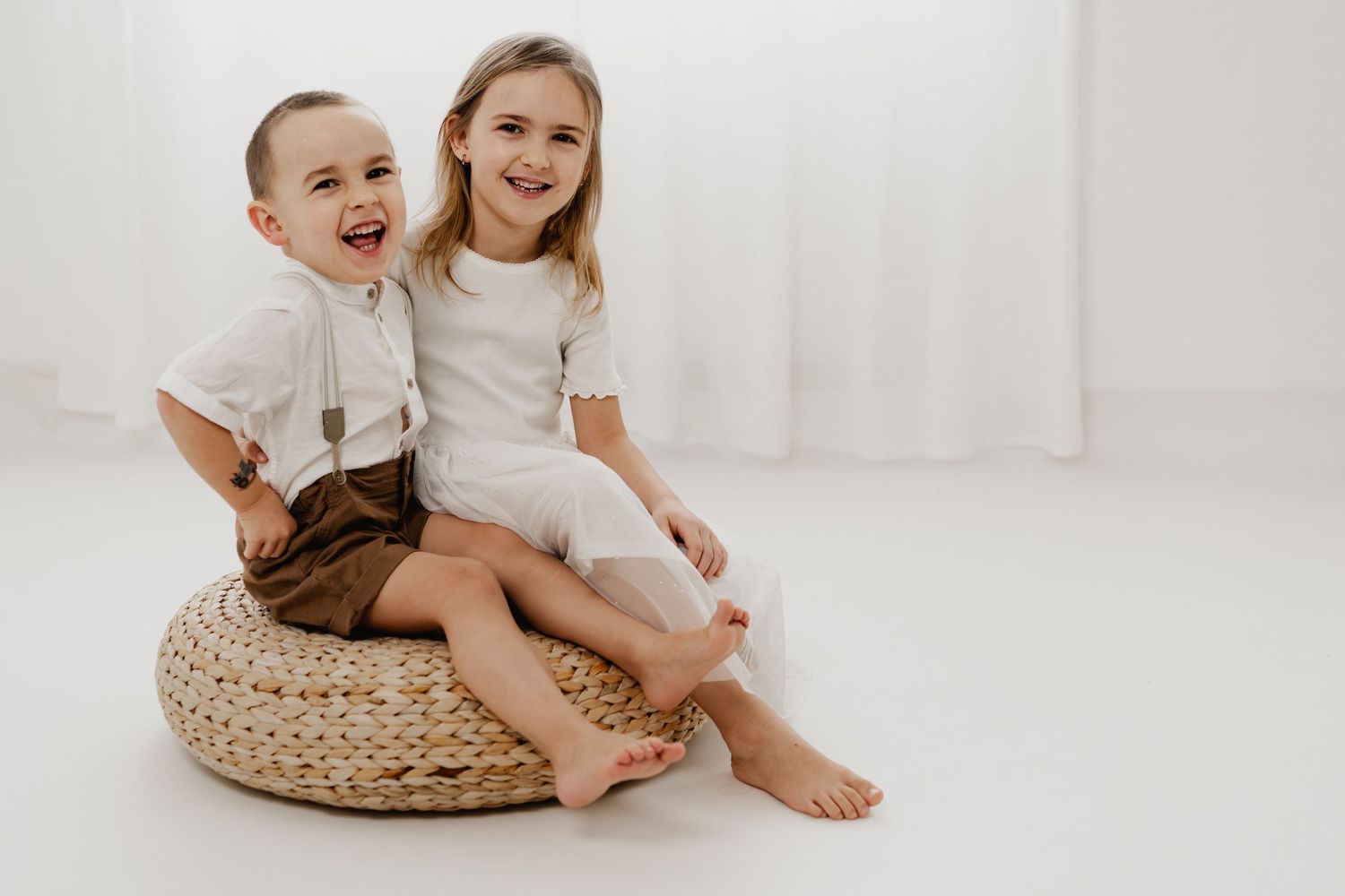 Bruder und Schwester sitzen lachend nebeneinander auf einem Korb-Hocker beim Familienfotoshooting im Fotostudio Stockerau