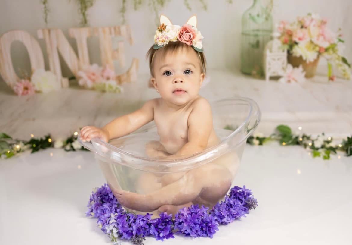 Baby in floral headband sitting in clear tub surrounded by purple flowers for first birthday photo.