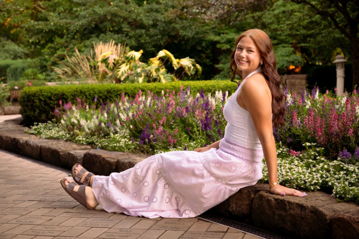 A person in a flowing pink dress poses gracefully in a garden setting with blooming flowers and greenery.