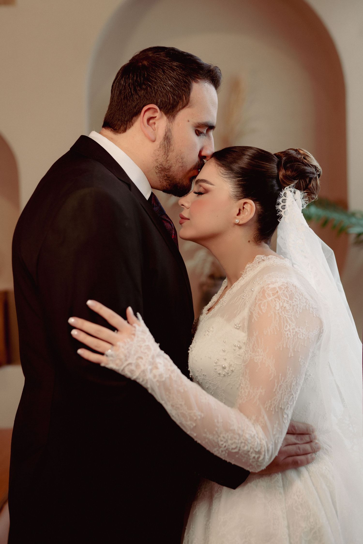 A tender moment between newlyweds as they embrace, with the bride wearing a lace-sleeved white gown and veil.