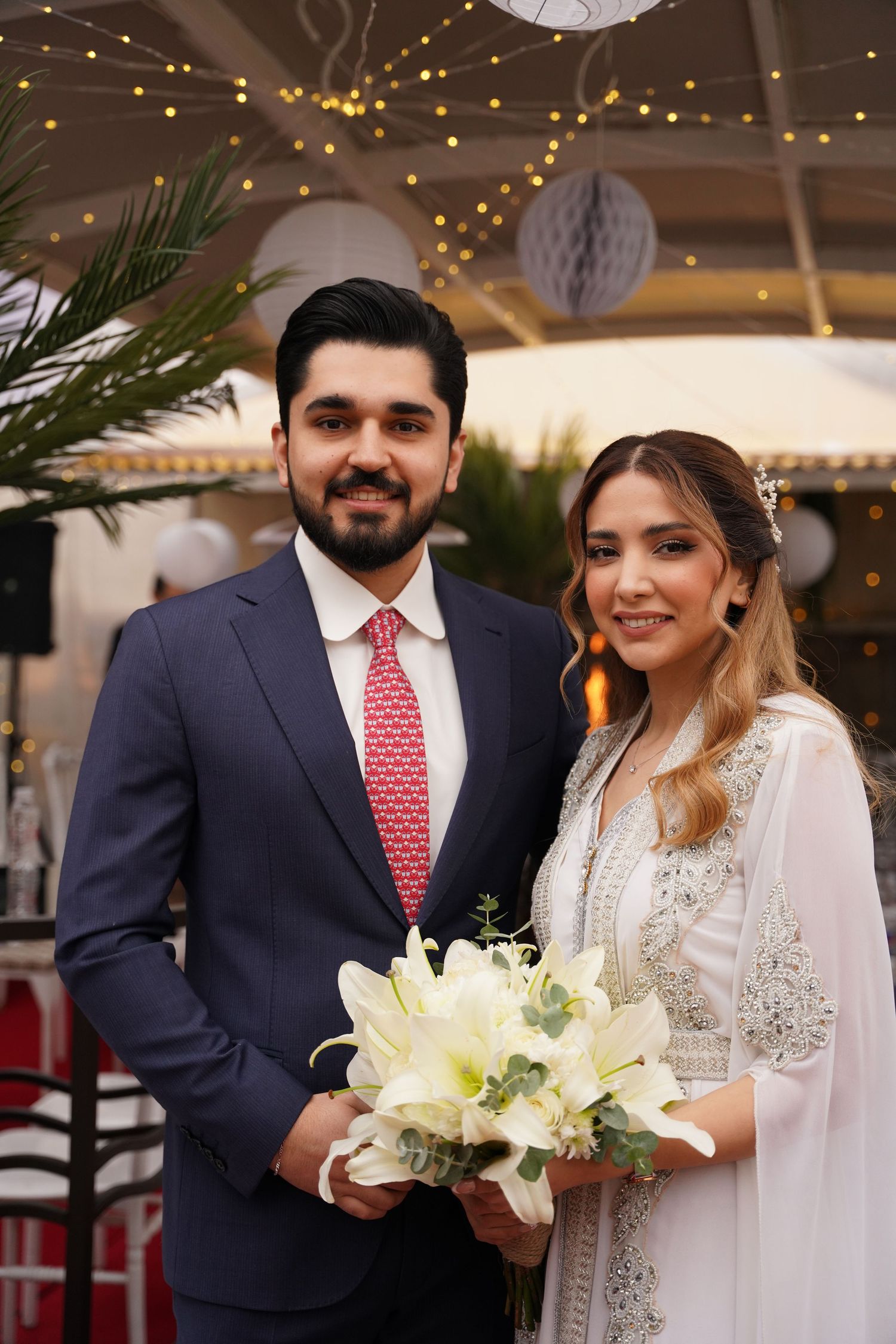 A couple at an elegant wedding celebration with string lights and decorations, bride holding a white lily bouquet.