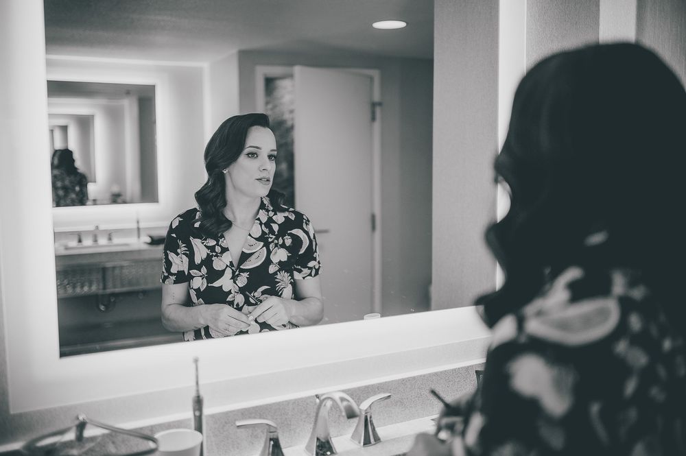 Black and white reflection in bathroom mirror shows person wearing floral print dress standing at vanity counter.