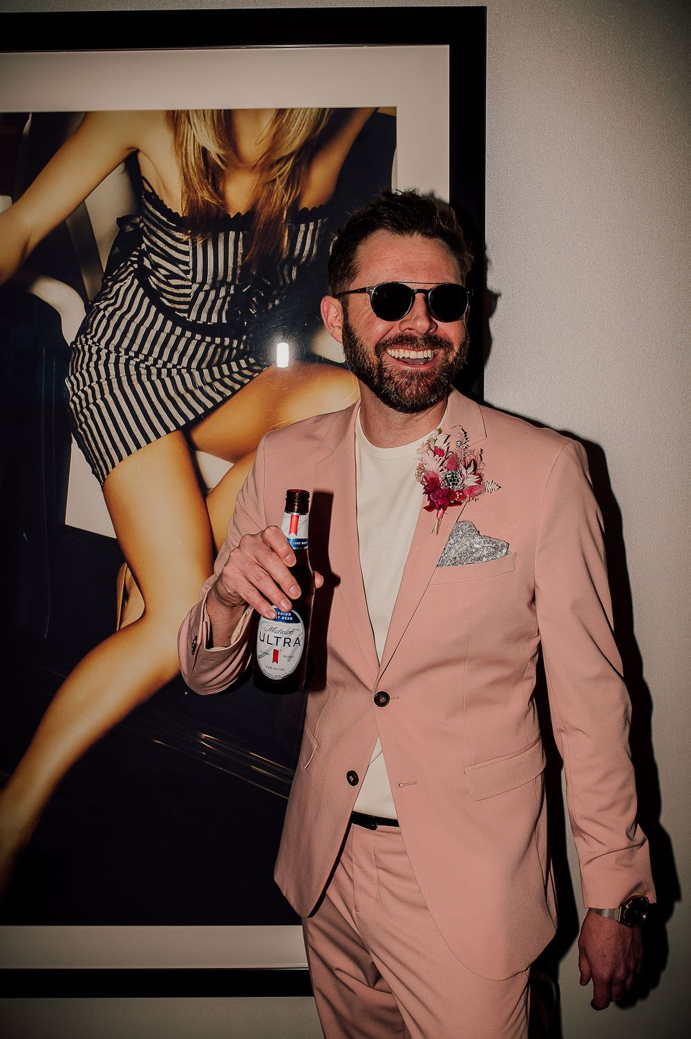 A person in a pink suit jacket and sunglasses poses at a party with a drink in their hand.