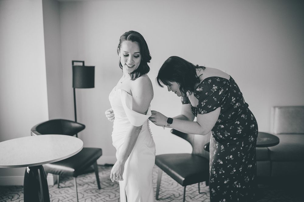 Black and white photo showing a person in a white dress being fitted by someone wearing a floral patterned dress.